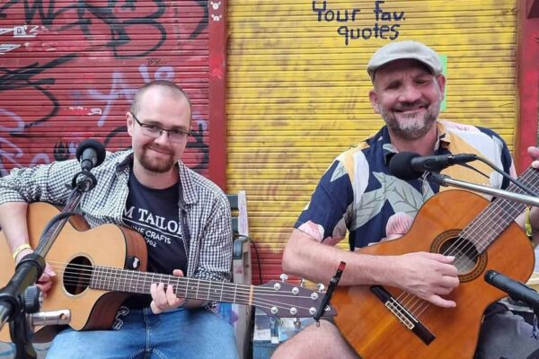 Buntfoto von zwei Männern mit Gitarren, sitzend nebeneinander mit dem Gesicht und Körper frontal zur Kamera, im Hintergrund deine bunte Wand