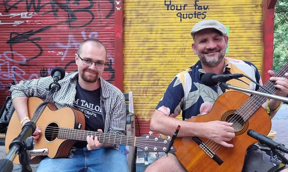 Buntfoto von zwei Männern mit Gitarren, sitzend nebeneinander mit dem Gesicht und Körper frontal zur Kamera, im Hintergrund deine bunte Wand