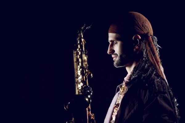 Buntfoto von Benjamin Michael im Profil vor einem dunklen Hintergrund mit einem Saxophon vor den geschlossenen Augen. Er trug eine dunkle Bandana und eine goldene Halskette mit dunklem Mantel.