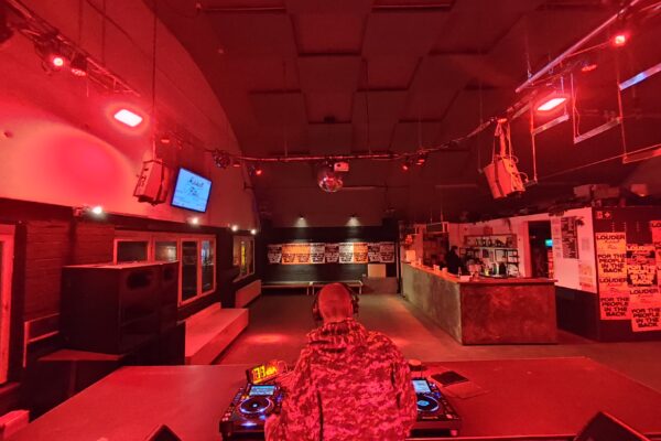 Buntfoto von einem Nachtclub, in rotes Licht getaucht. Im Vordergrund steht mit dem Rücken zur Kamera ein DJ und legt an zwei Plattenspieler und Technik auf, dahinter ein leerer Clubraum, geräumig mit Tanzfläche und langer Bar.