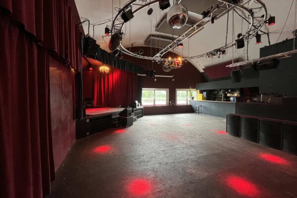 Buntfoto von einem Club, bei Tag, leer, mit Tanzfläche, Technikschienen darüber, roten Spotlights und einer Bühne im Hintergrund, auf der eine Band auftreten könnte, sowie einer Bar.