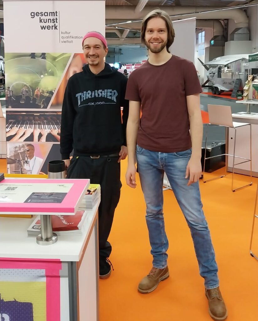 Buntfoto von zwei Männern, die auf einem Messtand stehen und in die Kamera lächeln. Im Hintergrund ist gesamtkunstwerk zu lesen, am unteren Bildrand ist die Ecke eines Pinc Music Plakats zu sehen.