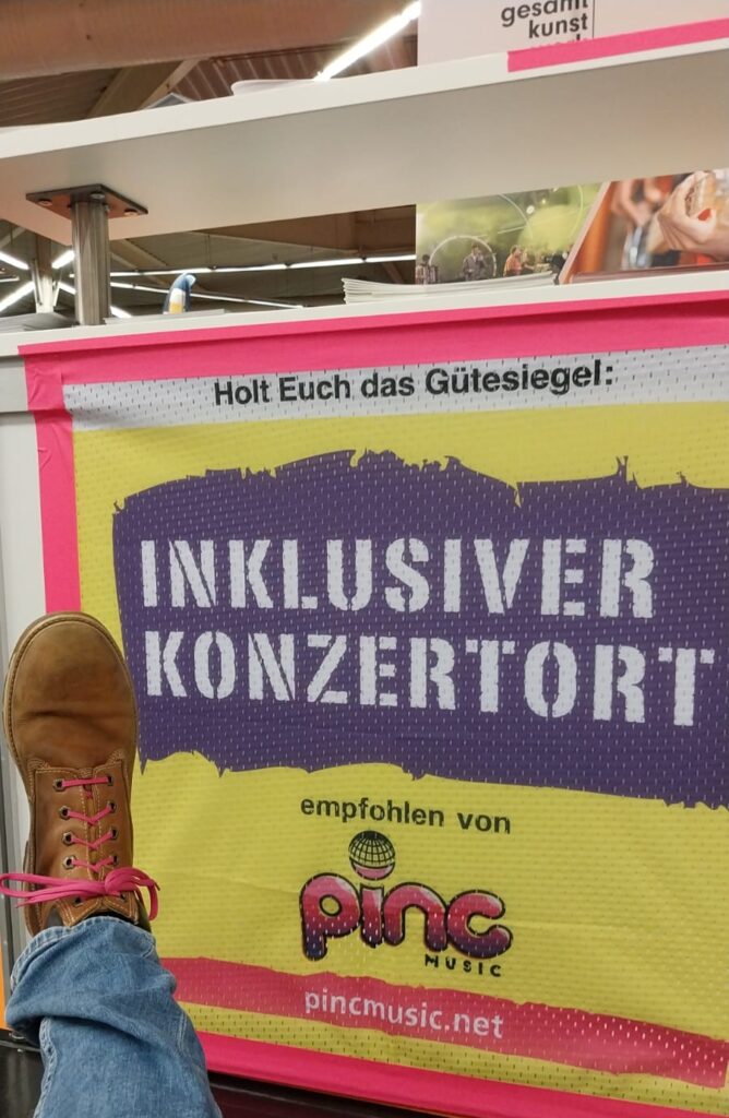 Buntfoto von einem Plakat mit Schrift "holt Euch das Gütesiegel: Inklusiver Konzertort empfohlen von Pinc Music" und davor ein ausgestrecktes Bein mit braunem Schuh, in den pinke Schnürsenkel eingezogen sind.