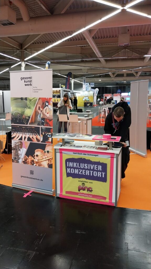 Buntfoto von einem Messestand, mit einem Tisch mit Pinc Music Plakat und einem Aussteller von gesamtkunstwerk, mit bunten Fotos von Musikern. Ein mann steht hinter dem Teisch und macht sich bereit für die Messe.