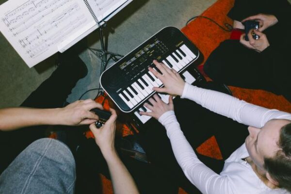 Buntfoto von einem Keyboard von oben fotografiert, eine Person spielt darauf, zwei weitere Personen stehen daneben und bedienen Joysticks.