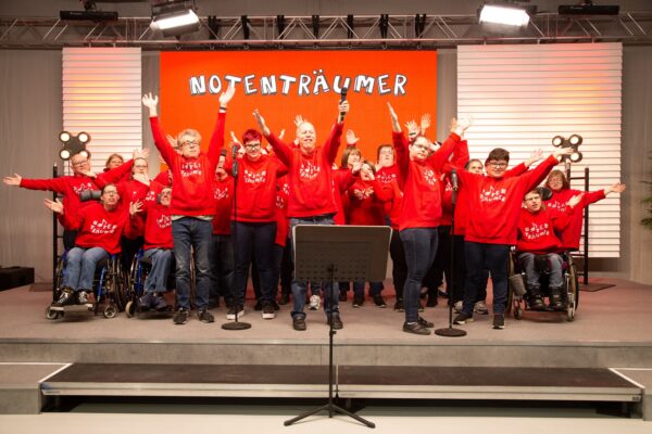 Buntfoto von einem Chor, mit über 20 Personen, auf einer Bühne stehend. Alle heben die Hände in die Luft. Im Hintergrund ein Banner mit der Anschrift "Notenträumer". Alle tragen das gleiche rote T-Shirt.