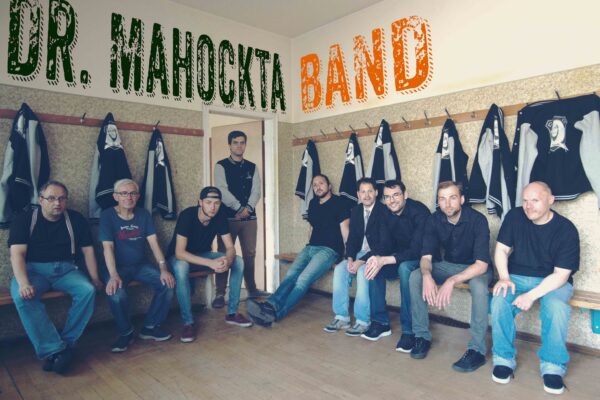 Auf dem Bild ist eine Gruppe von Menschen zu sehen, die als solche erkennbar ist, da sie augenscheinlich die gleichen Jacken besitzen, wo der Bandname "Dr. Mahockta" zu lesen ist. Diese Menschen sitzen in einer alten Sportumkleidekabine.