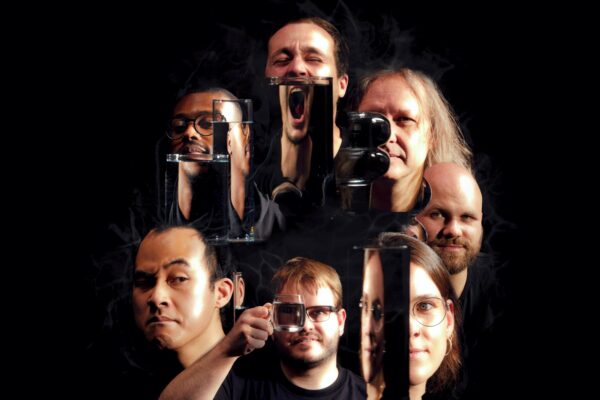 Ein buntes Portrait Gruppenfoto der Köpfe von sieben Menschen, die unterschiedliche Gesichter machen. Davor sind bei einigen mit einer durchsichtigen Flüssigkeit gefüllte Gläser zu sehen, die teils die Gesichter verdecken bzw. verfremden.