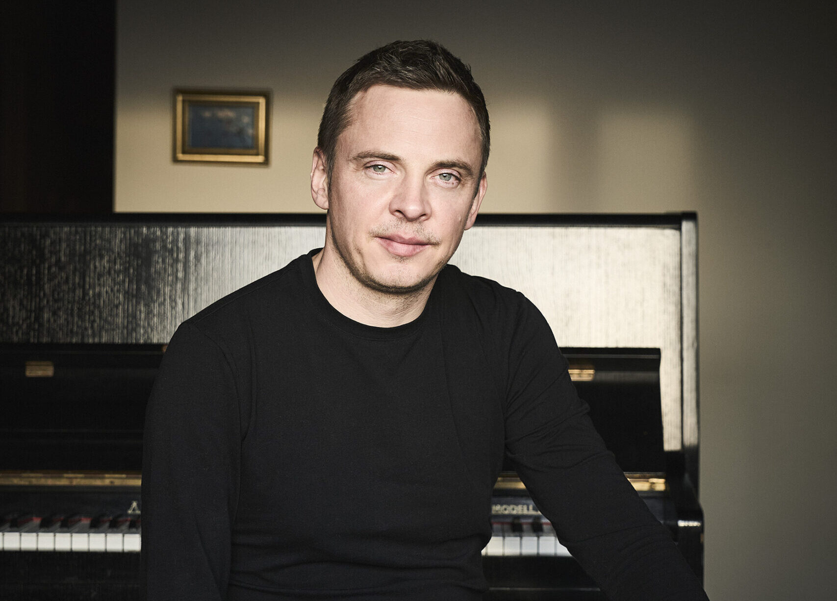 Buntfoto von einem Mann, sitzend. Er trägt einen schwarzen Pullover und guckt freundlichch in die Kamera. Er sitzt auf einem Hocker, vor einem Klavier.
