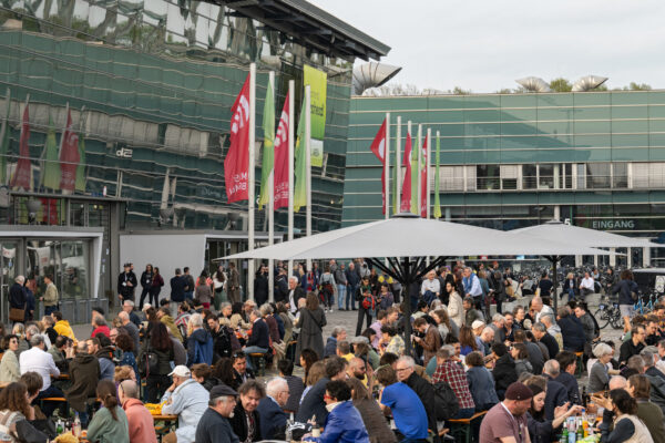 Buntfoto von einer Messehalle von außen mit fielen Menschen davor und Fahnenmasten mit Fahnen aus verschiedenen Ländern