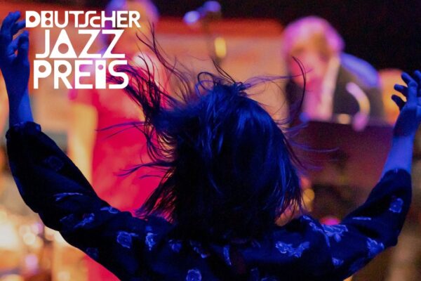 Eine Person tanzt mit erhobenen Armen auf einem Konzert. Ihr dunkles Haar ist in Bewegung, sie trägt ein Kleid mit Blumenmuster. Im Hintergrund erkennt man unscharf Musiker:innen auf der Bühne. Oben rechts steht das Logo des Deutschen Jazzpreises.