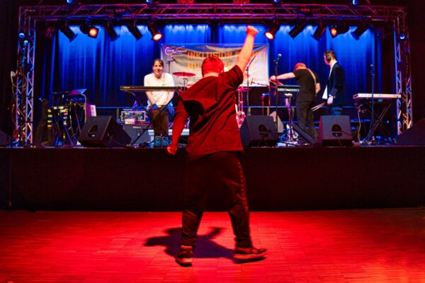 Buntfoto von einem Mensch. Er steht vor einer Bühne auf der eine Band Auftritt. Es ist innen und er ist in rotes Licht getaucht. Er steht da mit dem Rücken zur Kamera und nach oben erhobener rechten Faust.