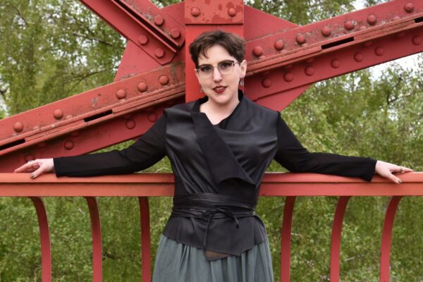 Farbfoto: Frau mit kurzen Haaren trägt Brillen, schwarzen Lederoptik-Top und grünen Rock steht mit Armen zu Seite ausgestreckt, auf einer roten Stahlbrücke angelehnt, und blickt lächelnd direkt in die Kamera, mit grünen Bäume im Hintergrund.