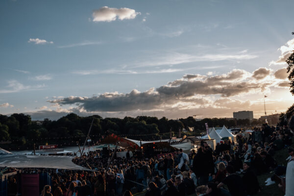 Buntfoto außen von einem Festivalgelände mit Zelten und Planen, vielen Menschen, darüber Himmel mit Wolken.