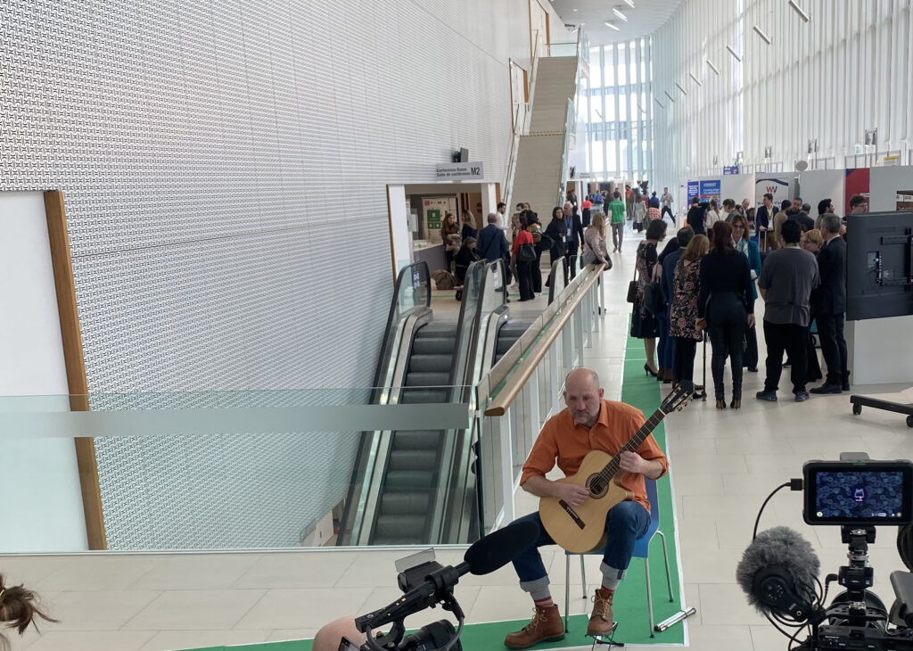 Buntfoto von einem Mann der auf einem Stuhl sitzt und Gitarre spielt. Im Vordergrund sind Mikrofone und ein mobiler monitor zu sehen, im Hintergrund eine Rolltreppe und ein Korridor mit vielen Menschen und Informationsständen.