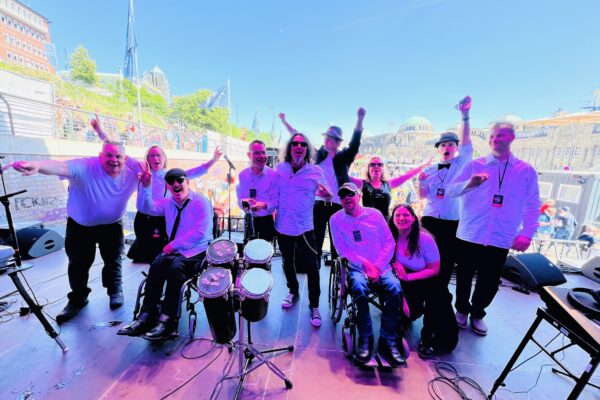 Verwaschenes Buntfoto von 11 Personen auf der Bühne, von hinter der Bühne fotografiert. Im Hintergrund ist der Platz vor den Landungsbrücken in Hamburg auszumachen und unscharf Publikum. Sechs der Personen heben die Arme, alle blicken freundlich bis euphorisch drein. 12 tragen weiße Hemden, eine ein schawarzts Kleid. Zwei Personen Sitzzen im Rollstuhl. Im Vordergrund stehen vier Bongos.