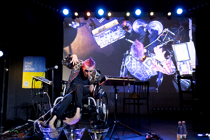 Buntfoto von einem Msuikperformer im Rollstuhl auf einer Bühne, im Hintergrund ist eine Lainwand auf der er von oben gefilmt live zu sehen ist. Neben ihm steht eine Loopstation. Seine nackten Füße sind in Schüsseln mit Wasser