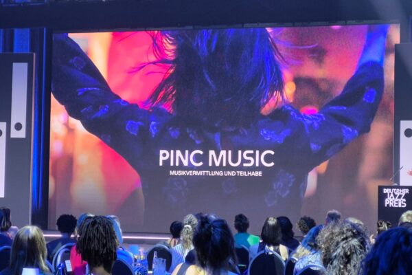 Buntfoto von großer Leinwand, darauf ein Bild von einer jungen Frau die vor einer Bühne jubelt (von hinten fotografiert) und dazu Schriftzug "Pinc Music" im Vordergrund Publikum.