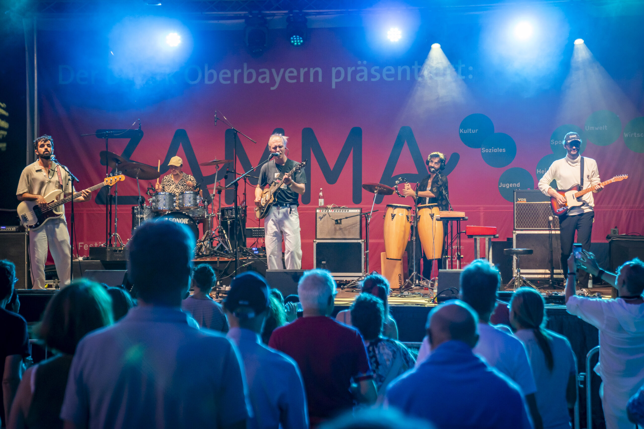Buntfoto von einer Bühne auf der fünf Musikerinnrnnen mit Schlagzeug, Gitarre, Bass und Congas auftreten. Im Hintergund Schriftzug "ZAMMA" im Verdergrund Publikum von hinten.