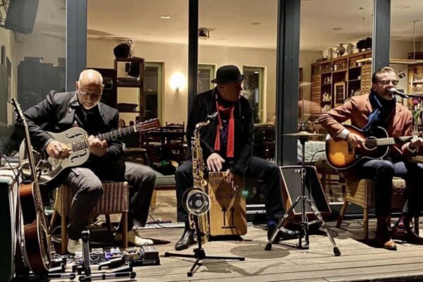 Buntfoto von drei Musikern die in Wohnzimmeratmoshpäre ein Konzert geben, mit Gitarre, Bass und Cajon