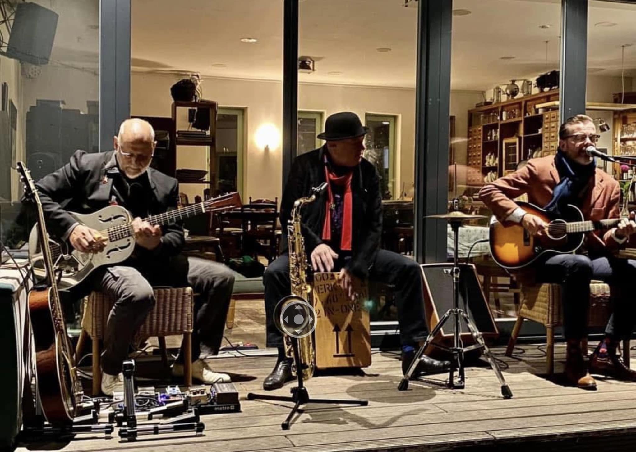Buntfoto von drei Musikern die in Wohnzimmeratmoshpäre ein Konzert geben, mit Gitarre, Bass und Cajon