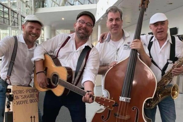 Buntfoto von vier Männern mit Cajon, Bass, Gitarre und Saxophon, alle sind gut gelauntund tragen weiße Hemden