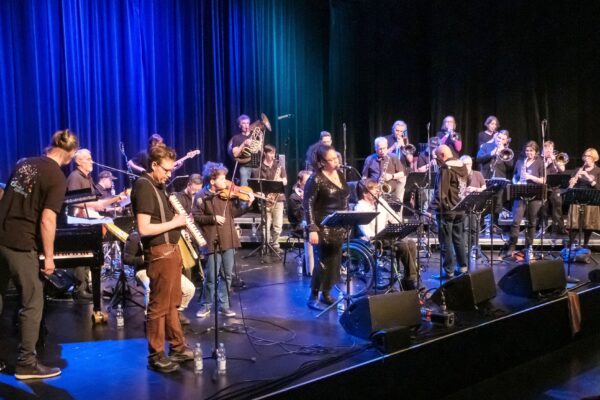 Buntfoto von einem weit über 20-köpfigen Orchester auf einer Bühne, mit vielen verschiedenen Instrumenten, im Hintergrund ein blauer Vorhang.