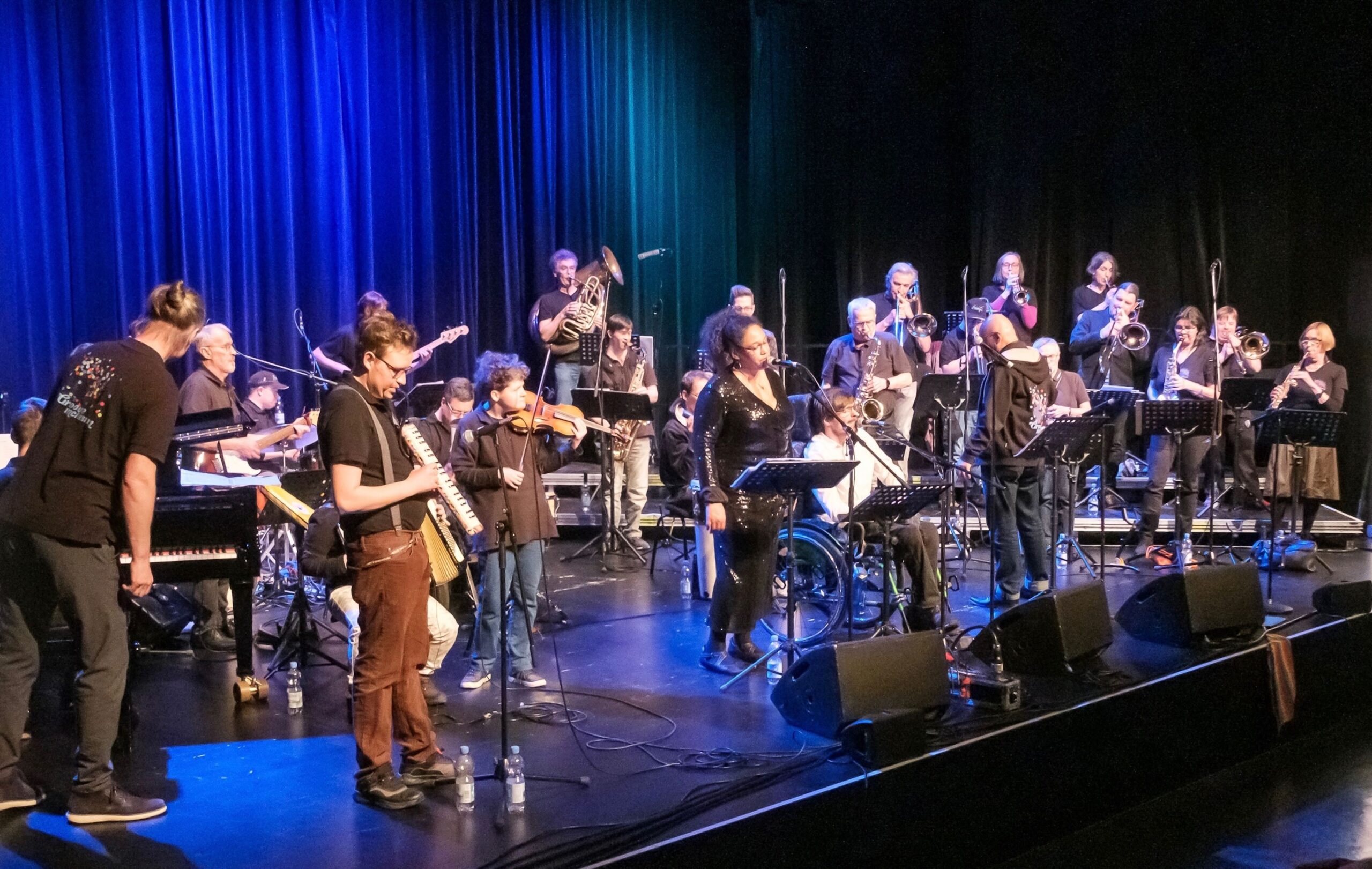 Buntfoto von einem weit über 20-köpfigen Orchester auf einer Bühne, mit vielen verschiedenen Instrumenten, im Hintergrund ein blauer Vorhang.
