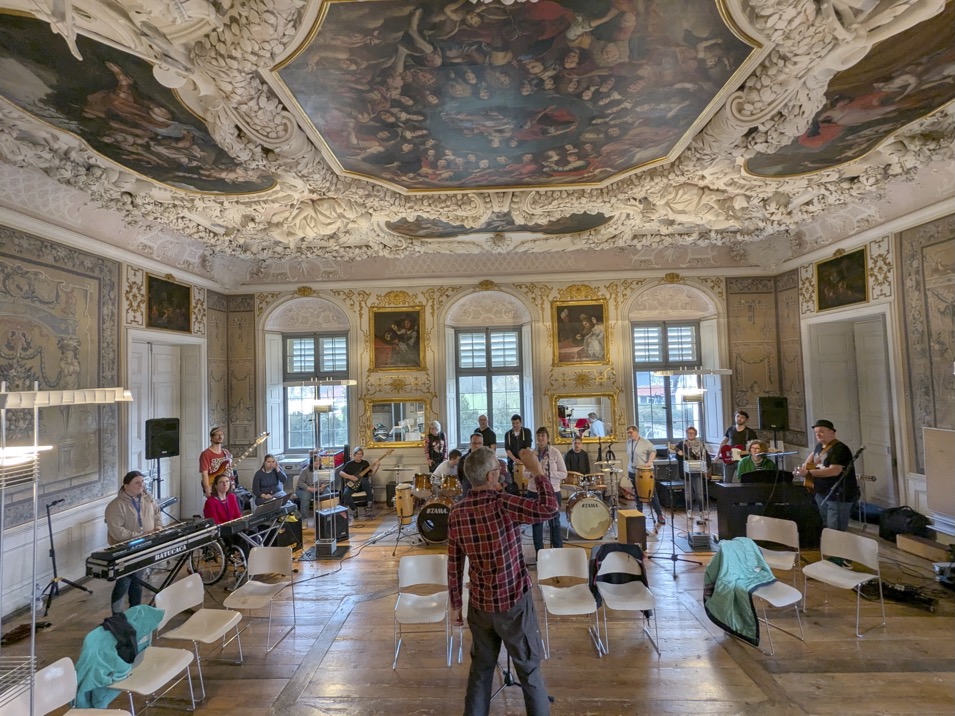 Buntfoto von einem prachtvollen Saal in einem Schloss, mit viel Stuck und großem buntem Decken-Gemälde, großen Fenstern, darin eine Band aus 18 Personen mit unterschiedlichsten Instrumendenten, die zusammen Proben. Ein Mann im Karrierten Hemd scheint zu dirigieren oder anzuleiten.