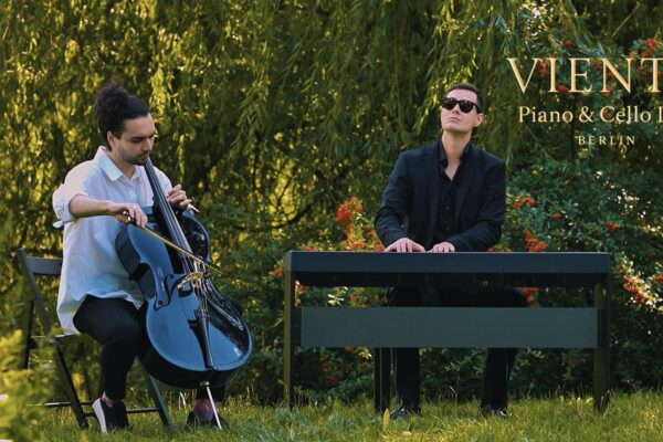 Buntfoto von zwei schick angezhogenen Männern (einer weißes Hemd, einer schwarzes Hemd und Jacket), sie sitzen in ienem grünen Park. Einer Spielt Chello, der andere Piano. Dzu Schrift: "VIENTOPiano & Cello Duo Berlin"