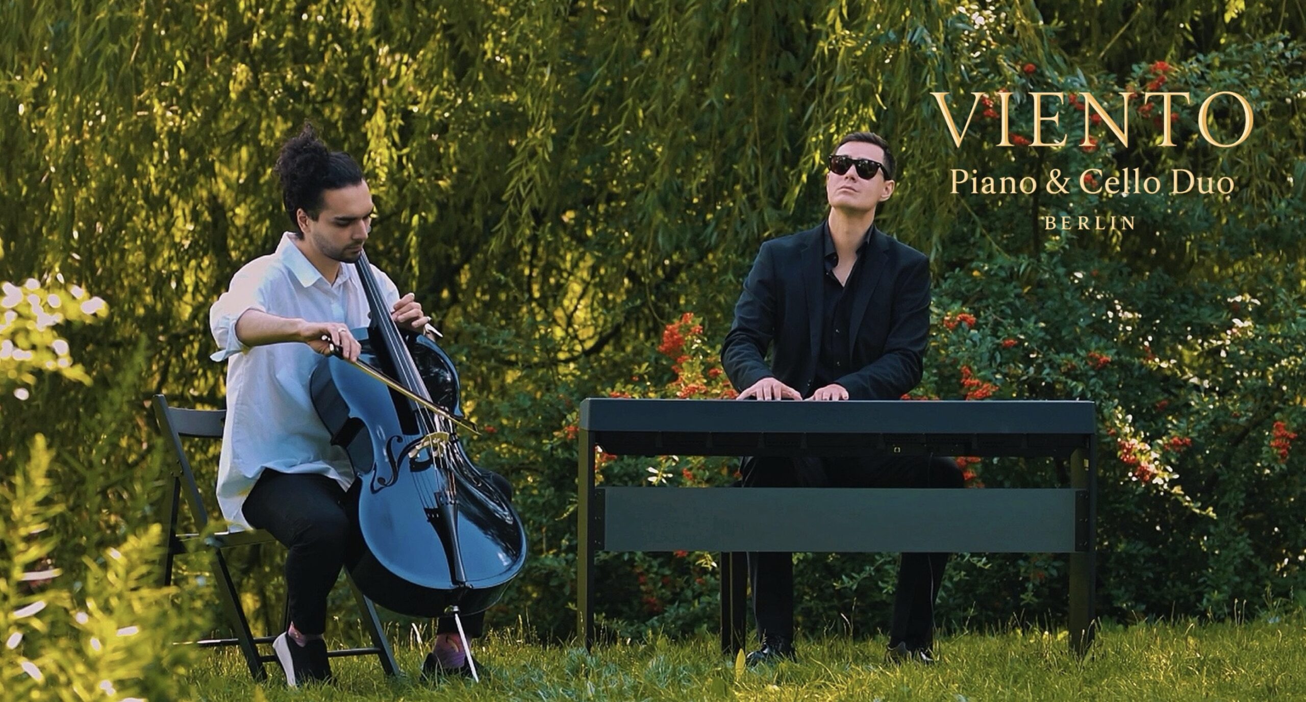 Buntfoto von zwei schick angezhogenen Männern (einer weißes Hemd, einer schwarzes Hemd und Jacket), sie sitzen in ienem grünen Park. Einer Spielt Chello, der andere Piano. Dzu Schrift: "VIENTOPiano & Cello Duo Berlin"