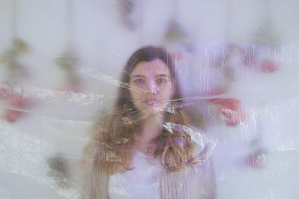 Buntfoto vom Kopf un Oberkörper einer Frau vor Rosen die an der Wand hängen. Sie und die Rosen sind hinter einer dicken transparenten Folie vor der Kamera verschwommen.