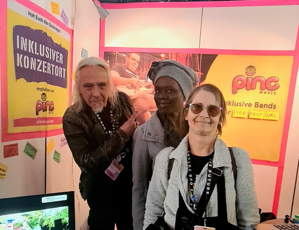 Buntfoto von drei gut gelaunen Menschen an einem Messestand, im Hintergrund ein Poster mit Schrift "Inklusiver Konzertort" und Pinc Music Logo