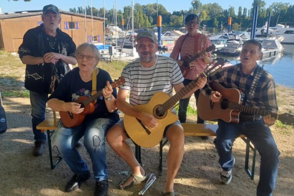 Buntfoto von einer Gruppe von fünf Menschen mit Gitarren und Ukulelen. Drei davon sitzen nebeneinander auf einer Bierbank, die anderen beiden stehen dahinter. Alle gucken freundlich in die Kamera. Im Hintergrund liegt ein Hafen mit kleinen Segelbooten.
