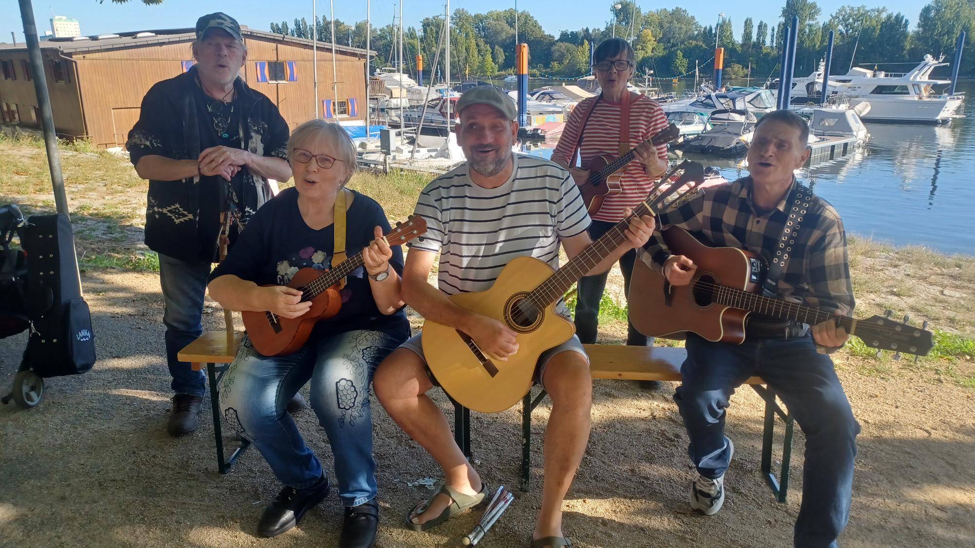 Buntfoto von einer Gruppe von fünf Menschen mit Gitarren und Ukulelen. Drei davon sitzen nebeneinander auf einer Bierbank, die anderen beiden stehen dahinter. Alle gucken freundlich in die Kamera. Im Hintergrund liegt ein Hafen mit kleinen Segelbooten.