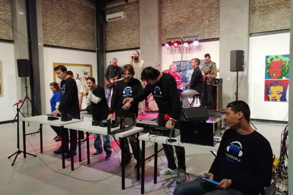 Buntfoto von ienem Orchester von 12 Menschen mit verschiedenen Instrumenten (Schlagzeug, E-Gitarre, E-Bass, Keyboard) und verschiedener Technik (Mihscpult, Tablets) live bei einem Auftritt. 5 Davon sitezen/stehen auf einer kleinen erhöhten Bühne, die anderen stehen davor hinter Tischen, auf denen TEchnik aufgebaut ist.