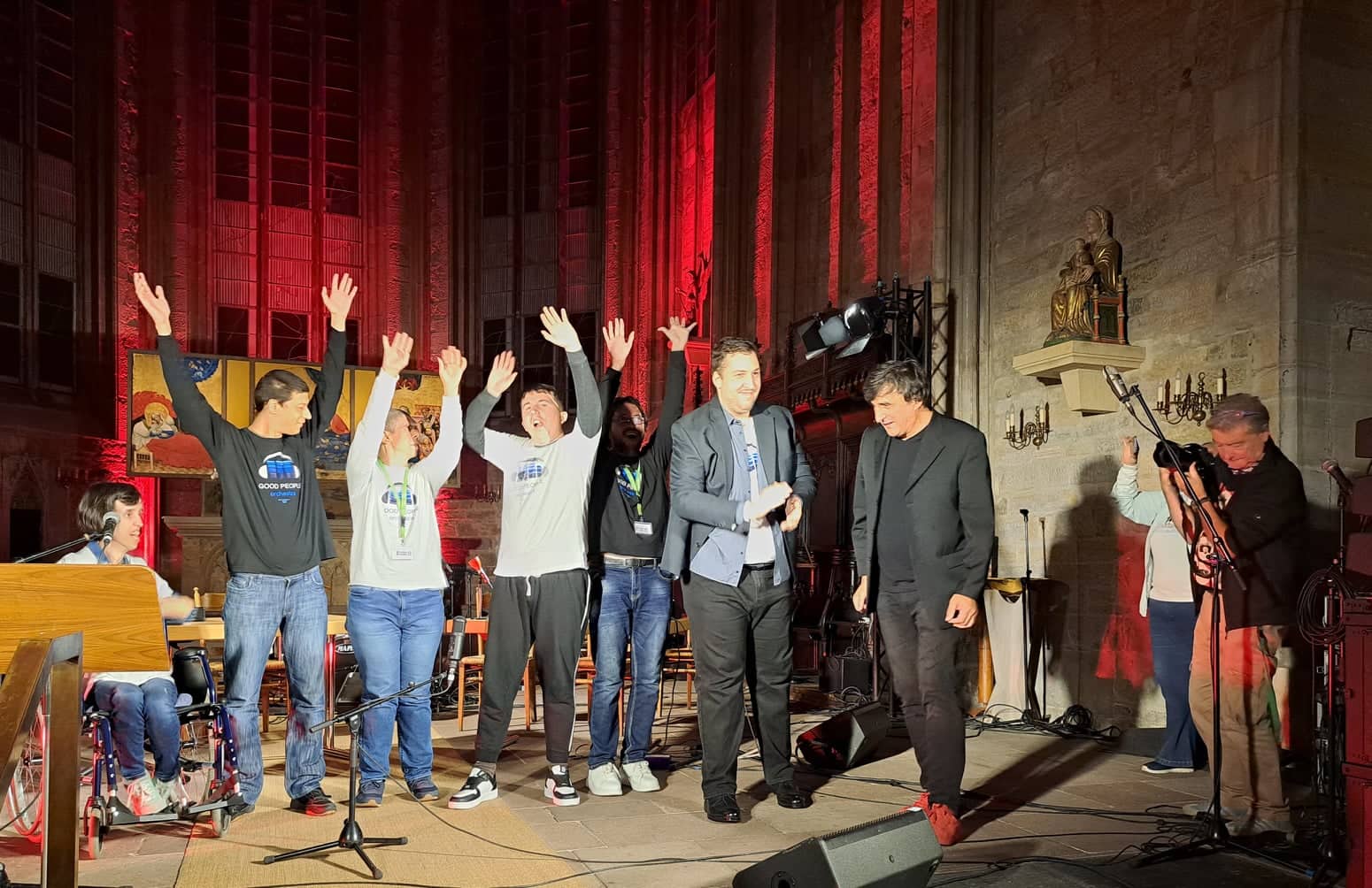 Buntfoto von 7 Menschen nebeinander einer Reihe, einer sittz im Rollstuhl, der rest Steht, davon recken fünf die Arme und Hände hoch in die Luft und einer klatsch. Im Hintergrund ein Altar und die Hohen Fenster und Wände einer Kirche.
