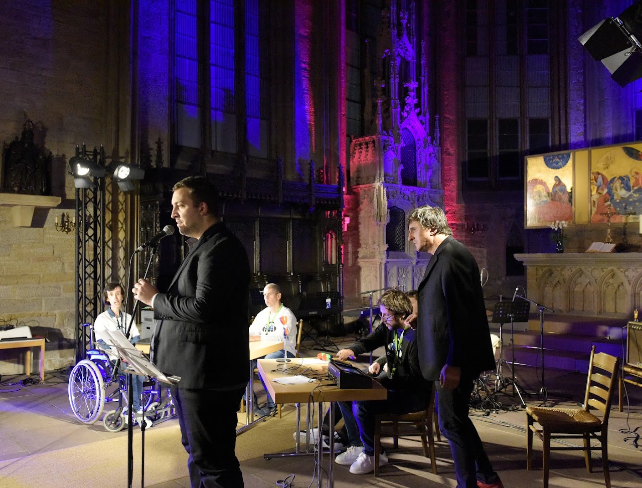 Buntfoto von sechs Menschen in einer Kirche. Einer steht an einem Mikrofon im Ständer und singt, einer sitzt an einem Mischpult, einer sitzt im Rollstuhl und zwei sind im Anschnitt, einer steht. Im HIntergrund ein Altar und verzierte Wände der Kirche