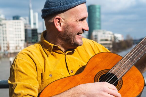 Buntfoto von einem Mann. Er steht vor einem Geländer vor einem Fluss, im Hintergrund auf der anderen Seite des Flusses sind Hochhäuser. Er trägt ein gelbes Hemd und eine blaue Wollmütze und spielt Gitarre.
