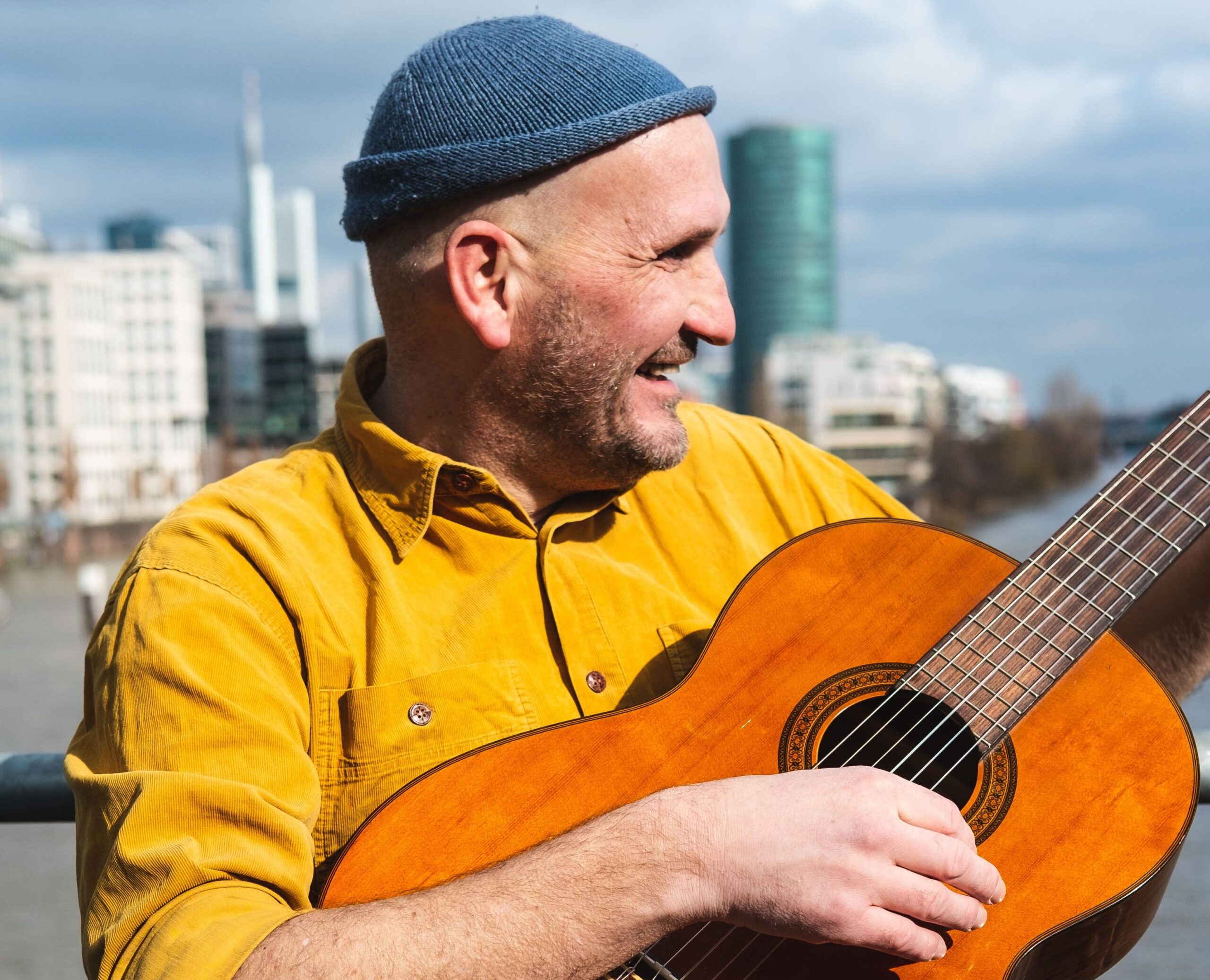 Buntfoto von einem Mann. Er steht vor einem Geländer vor einem Fluss, im Hintergrund auf der anderen Seite des Flusses sind Hochhäuser. Er trägt ein gelbes Hemd und eine blaue Wollmütze und spielt Gitarre.