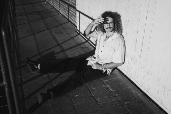 Das Bild ist in schwarz-weiß. Calvin sitzt angelehnt an einer Wand. Seine Beine sind ausgestreckt. Seine rechte Hand schirmt leicht die Sonne ab und wirft einen kleinen Schatten auf sein Gesicht. Calvin schaut nach links.