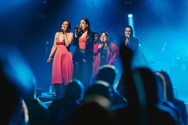 Buntfoto von vier freuen, die im blauen Scheinwerferlicht auf einer Bühne stehen und alle in ihr Mikrofon singen. Sie sind festlich gekleitet.