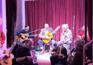 Mehrere Musikerinnen sitzen auf einer kleinen Bühne vor bordeauxroten Vorhängen. Drei Personen spielen Akustikgitarren, eine weitere ein kleines Saiteninstrument. Im Vordergrund des Publikums spielen zwei Personen ebenfalls Instrumente, darunter eine Melodica und eine kleine Gitarre. Die Szene ist warm ausgeleuchtet und wirkt lebendig und gemeinschaftlich.*