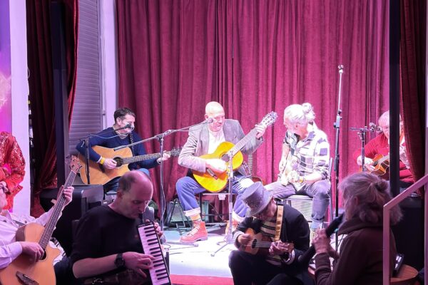 Mehrere Musikerinnen sitzen auf einer kleinen Bühne vor bordeauxroten Vorhängen. Drei Personen spielen Akustikgitarren, eine weitere ein kleines Saiteninstrument. Im Vordergrund des Publikums spielen zwei Personen ebenfalls Instrumente, darunter eine Melodica und eine kleine Gitarre. Die Szene ist warm ausgeleuchtet und wirkt lebendig und gemeinschaftlich.*