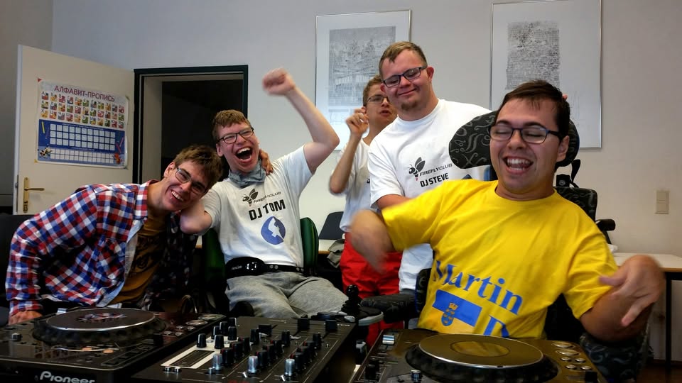 Mehrere fröhliche Firefly-Club DJs sitzen gemeinsam an einem DJ-Pult, lachen, jubeln und genießen sichtbar den Moment. Die Szene strahlt Energie, Freude und Teamgeist aus.