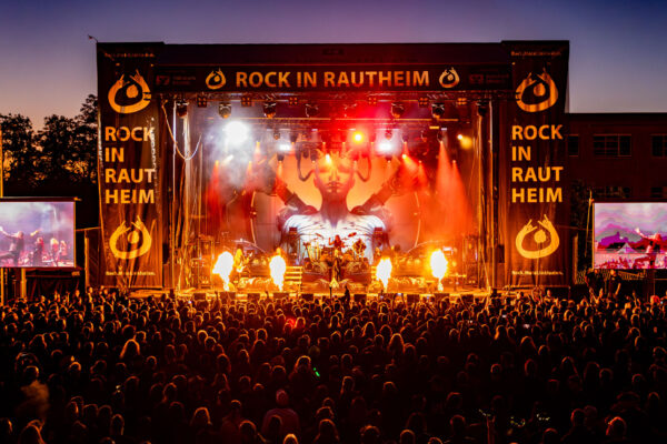 Buntfoto. Große Open-Air-Bühne in hellem roten und orangefarbenen Licht, Effekte von Flammen auf der Bühne, auf der Bühne eine Band. Im Hintergrund ist eine übergroße Figur als Wesen zwischen Mensch und künstlichem Wesen projiziert. Eine große Menschenmenge vor der Bühne.