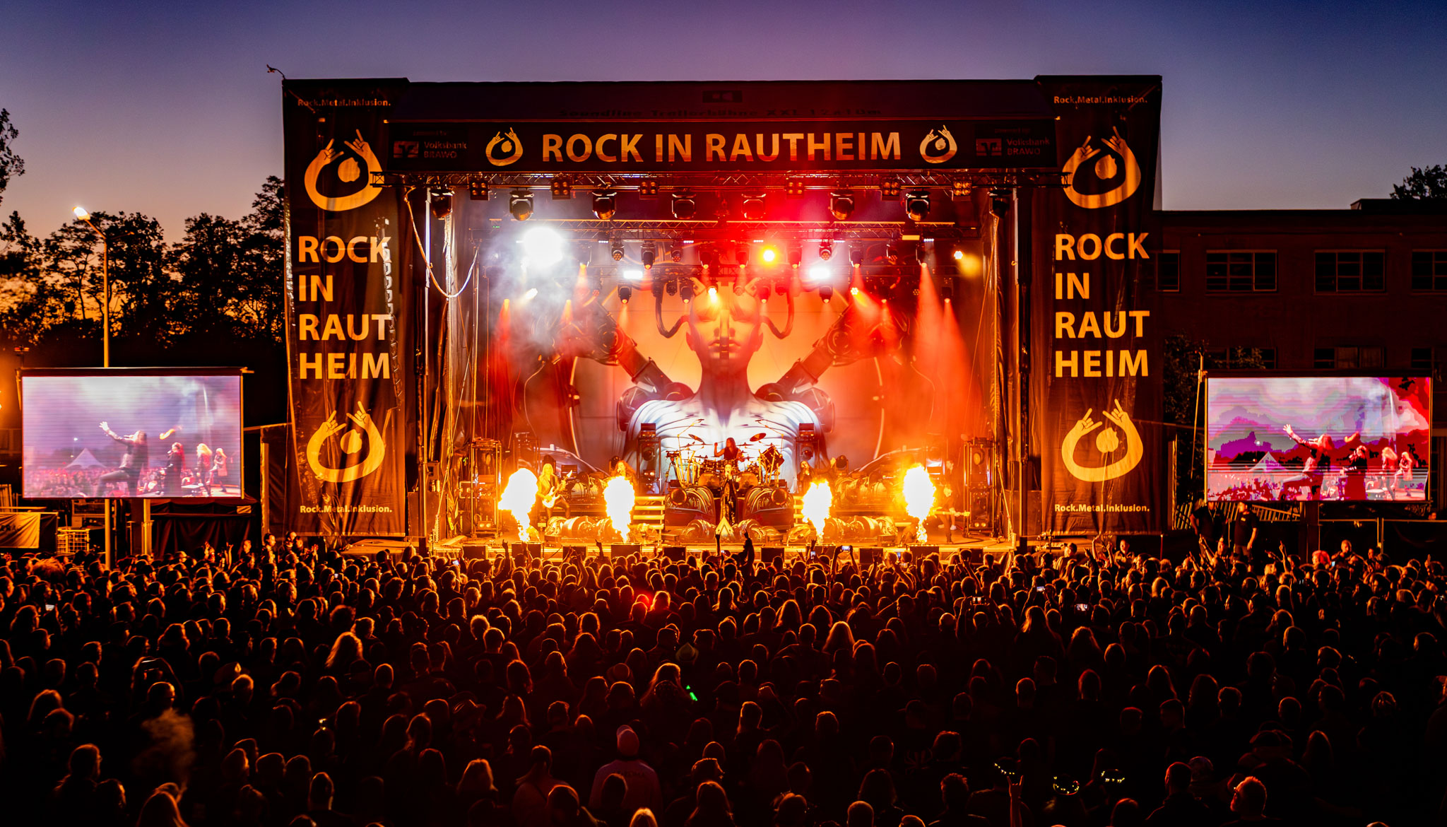 Buntfoto. Große Open-Air-Bühne in hellem roten und orangefarbenen Licht, Effekte von Flammen auf der Bühne, auf der Bühne eine Band. Im Hintergrund ist eine übergroße Figur als Wesen zwischen Mensch und künstlichem Wesen projiziert. Eine große Menschenmenge vor der Bühne.
