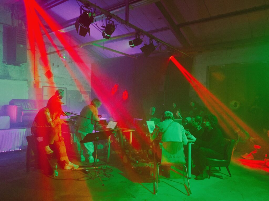 Mehrere Menschen machen Musik in einem dunklen Raum. Buntes Licht in Rot und Grün leuchtet auf die Bühne. Die Musiker sitzen und stehen im Kreis. Instrumente und Technik stehen im Raum. Auch Zuhörer*innen sind dabei. Das Bild zeigt eine experimentelle Musik-Situation mit Live-Klängen.