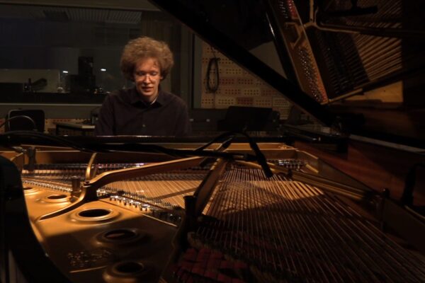 Buntfoto von einem jungen Mann an einem Flügel, durch den aufgeklappten Deckel des Flügels hindurch fotografiert.