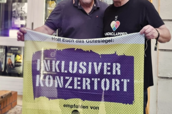 Bildbeschreibung: Buntfoto von zwei Männern die vor dem Eingang einer Kleinkunstbühne stehen, an dem einige Plakate hängen. Sie halten ein großes Stoffbanner hoch auf dem steht "holt Euch das Gütesiegel: Inklusiver Konzertort empfohlen von Pinc Music. pincmusic.net"
